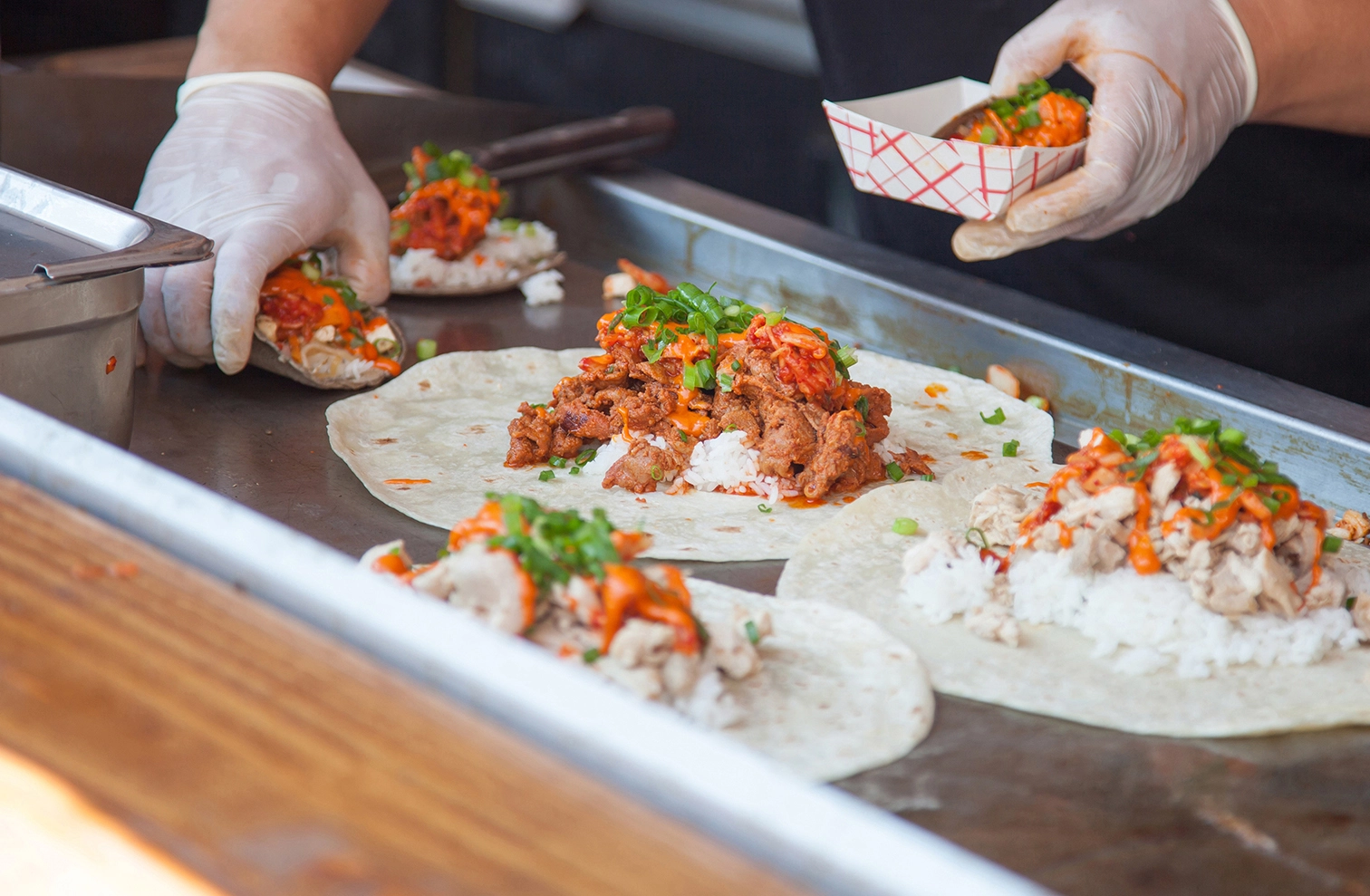 Gloved hands assembling tacos on a griddle with meat, rice, and salsa.