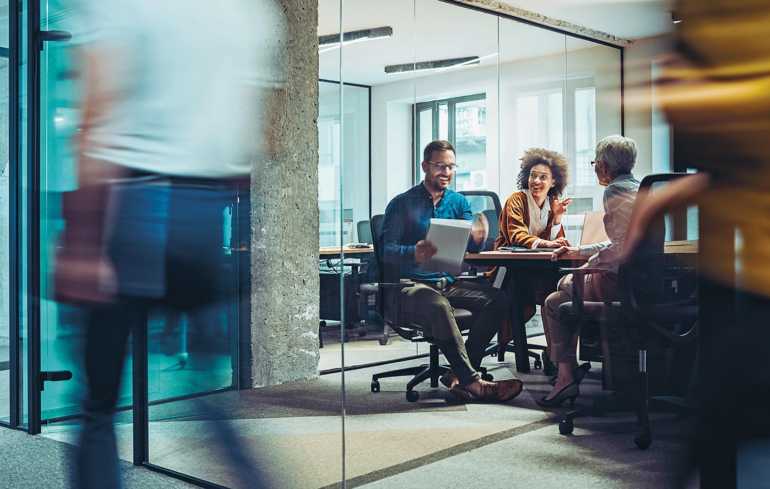 Colleagues meeting in a glass-walled office as people walk by.